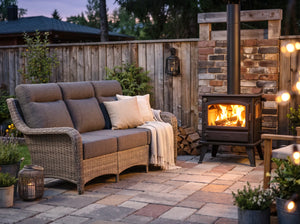 OuterCraft 3-seater wicker sofa with dark gray cushions showcased in an evening patio setting with wood stove.