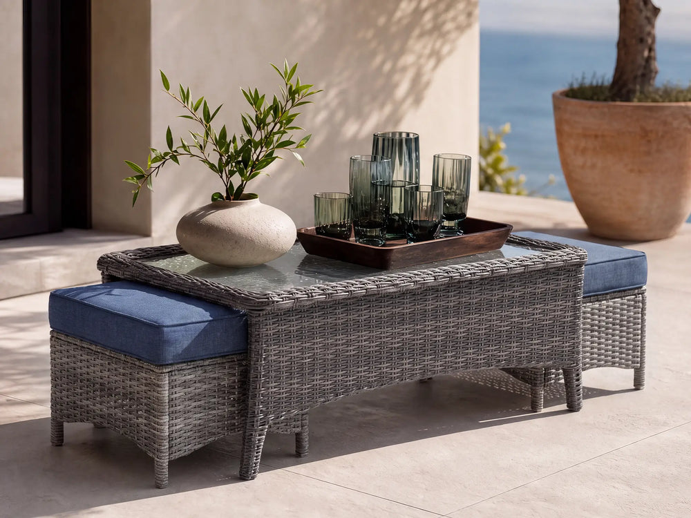 OuterCraft Monolith outdoor coffee table featuring an inset tempered glass top and two space-saving nesting ottomans with blue cushions, crafted in hand-woven dark all-weather resin wicker on a luxury coastal terrace.